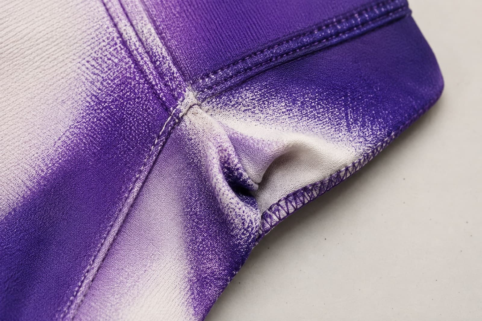 Macro photograph of a printed polyester seam where the violet colour thins to fine white speckling along the stitch line and inside the fold.